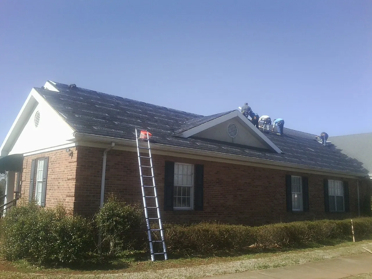 Crew tearing shingles off a brick ranch roof with a ladder set — Emergency Roof Repair work by our crew in Bridgehampton